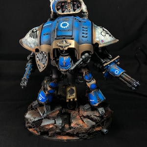 K&ouml;nnte beinhalten: Ein blauer und goldfarbener bemalter Miniaturroboter mit einer gro&szlig;en Waffe, der auf einem felsigen Sockel steht. Der Roboter hat einen gro&szlig;en Kopf mit einer wei&szlig;en Feder und eine gro&szlig;e Waffe am rechten Arm.
