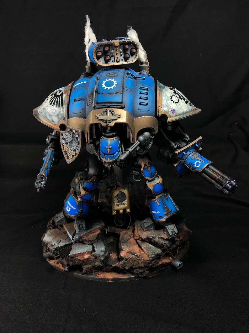 K&ouml;nnte beinhalten: Ein blauer und goldfarbener bemalter Miniaturroboter mit einer gro&szlig;en Waffe, der auf einem felsigen Sockel steht. Der Roboter hat einen gro&szlig;en Kopf mit einer wei&szlig;en Feder und eine gro&szlig;e Waffe am rechten Arm.