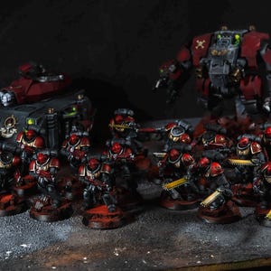 Op de afbeelding: Miniatuurfiguren van een sci-fi leger, met soldaten in rode en zwarte bepantsering, een rode tank en een grote robot. De figuren staan op een donkere ondergrond, met een donkere achtergrond. De miniaturen zijn gedetailleerd.