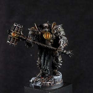 May include: A miniature figurine of a heavily armored warrior with a large hammer. The figure is painted in metallic grey and bronze, with gold highlights. The base is textured to resemble rocky terrain. The overall aesthetic suggests a fantasy theme, suitable for tabletop gaming.