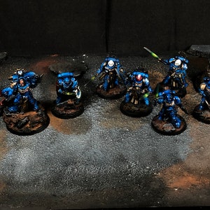 K&ouml;nnte beinhalten: Eine Gruppe von blau und silber bemalten Miniaturfiguren, die auf runden Sockeln stehen, mit Schwertern und anderen Waffen. Die Figuren sind in einer Reihe angeordnet, mit einigen Figuren in der hinteren Reihe.