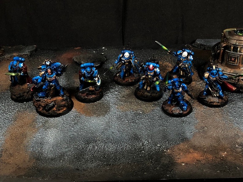 K&ouml;nnte beinhalten: Eine Gruppe von blau und silber bemalten Miniaturfiguren, die auf runden Sockeln stehen, mit Schwertern und anderen Waffen. Die Figuren sind in einer Reihe angeordnet, mit einigen Figuren in der hinteren Reihe.