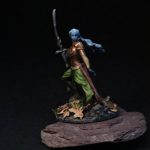 May include: A blue-skinned miniature figurine of a female warrior holding a sword, standing on a base with brown leaves and a dark gray rock.