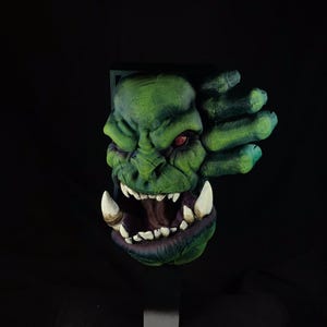 May include: A green and purple Orc head sculpture with a large open mouth and sharp teeth. The sculpture is mounted on a black stand.