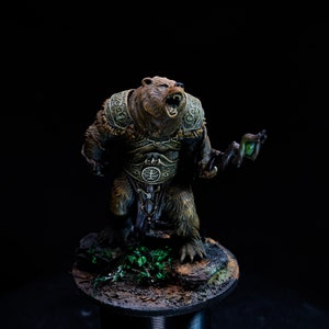 Puede incluir: Una figura miniatura pintada de un oso con armadura y sosteniendo un bastón. El oso es marrón y tiene un bastón verde con un orbe brillante en el extremo. El oso está de pie sobre una base con vegetación verde y tierra marrón.