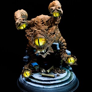 Peut inclure: Figurine détaillée d'une créature fantastique marron avec de multiples yeux jaunes et des dents pointues. La figurine est posée sur une plateforme en pierre avec des livres et d'autres objets. Le fond est noir.