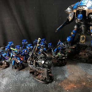 K&ouml;nnte beinhalten: Ein blaues und goldenes bemaltes Miniatur-Rittermodell steht hoch aufragend &uuml;ber einer Gruppe kleinerer, blau bemalter Miniaturfiguren. Der Ritter h&auml;lt ein gro&szlig;es Schwert und einen Schild, w&auml;hrend die kleineren Figuren mit verschiedenen Waffen bewaffnet sind. Die Figuren stehen alle auf kleinen Sockeln, und der Ritter steht auf einem gr&ouml;&szlig;eren Sockel.