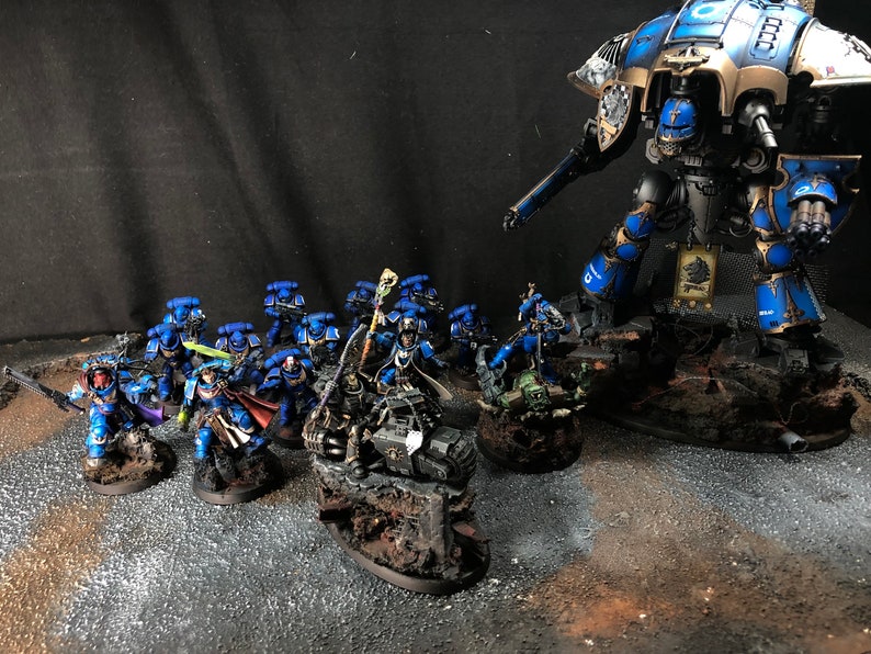 K&ouml;nnte beinhalten: Ein blaues und goldenes bemaltes Miniatur-Rittermodell steht hoch aufragend &uuml;ber einer Gruppe kleinerer, blau bemalter Miniaturfiguren. Der Ritter h&auml;lt ein gro&szlig;es Schwert und einen Schild, w&auml;hrend die kleineren Figuren mit verschiedenen Waffen bewaffnet sind. Die Figuren stehen alle auf kleinen Sockeln, und der Ritter steht auf einem gr&ouml;&szlig;eren Sockel.