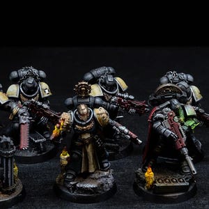 May include: A collection of miniature figurines of armored soldiers, painted in black, tan, and red. The figures are posed in various action stances, some holding weapons. The bases are detailed with a textured, rocky appearance, and the background is dark.