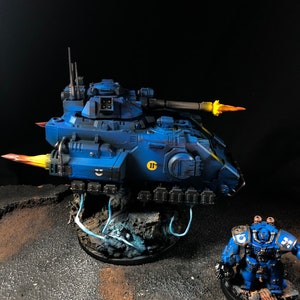 K&ouml;nnte beinhalten: Ein blauer und gelber bemalter Miniaturpanzer mit einem blauen und gelben bemalten Miniatur-Space Marine, der daneben steht. Der Panzer hat eine gro&szlig;e Kanone und feuert eine gelbe Flamme ab. Der Space Marine h&auml;lt eine Waffe und tr&auml;gt einen blauen Helm.