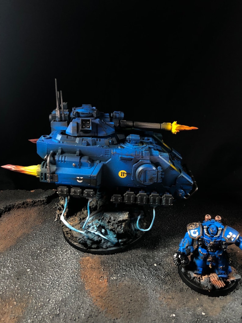 K&ouml;nnte beinhalten: Ein blauer und gelber bemalter Miniaturpanzer mit einem blauen und gelben bemalten Miniatur-Space Marine, der daneben steht. Der Panzer hat eine gro&szlig;e Kanone und feuert eine gelbe Flamme ab. Der Space Marine h&auml;lt eine Waffe und tr&auml;gt einen blauen Helm.