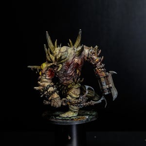 Könnte beinhalten: Eine bemalte Miniaturfigur einer massigen, monströsen Kreatur mit blasser, texturierter Haut, scharfen, hornartigen Vorsprüngen an den Schultern und einer großen Metallklinge am linken Arm, die eine gekettete Waffe in der rechten Hand hält.