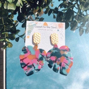 May include: A pair of handmade earrings featuring a tropical leaf design with pink, blue, orange, and black paint. The earrings are attached to gold-coloured, textured, oval-shaped posts. The card reads "Sweet to the Soul Handmade earrings".