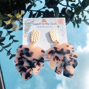 Puede incluir: Un par de pendientes en forma de hoja con un estampado carey en tonos negro, marrón y crema. Los pendientes tienen la parte superior texturizada de color dorado y se muestran en una tarjeta que dice "Sweet to the Soul handmade earrings."