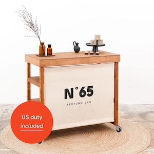 Può includere: Tavolo espositivo in legno per un laboratorio di profumi, con un fronte in tessuto color crema con la scritta "N°65 PERFUME LAB" in nero. Il tavolo ha un ripiano, ruote ed è decorato con bottiglie di profumo, una piccola brocca nera e saponi.