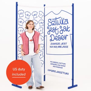 May include: A woman stands in front of a white banner with blue accents and text in Polish. The banner reads "Sztuka jest jak Deser" and features a t-shirt design. The woman wears a pink vest over a white t-shirt and light blue jeans.