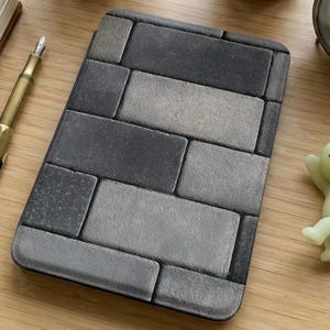 May include: A rectangular case with a brick pattern in shades of gray. The case is made of a textured material and has rounded corners. The design resembles a brick wall. The case is on a wooden surface.