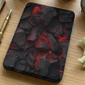 Può includere: Custodia rettangolare nera e rossa con un motivo a roccia vulcanica. La custodia, probabilmente per un tablet o un e-reader, è appoggiata su una superficie di legno. Sono visibili anche una penna e una piccola statuina.
