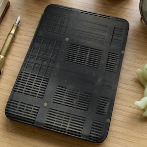 May include: A black laptop base with a grid-like pattern of vents and rectangular sections. The base is rectangular with rounded corners and is lying on a wooden surface. A gold pen is visible in the background.