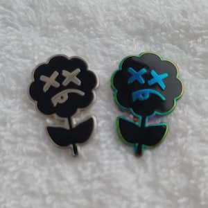 May include: Two flower-shaped enamel pins with black petals and stems. One pin has a silver outline and white "XX" eyes, the other has a rainbow outline and blue "XX" eyes. The pins are on a white, textured surface.