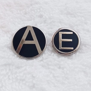 May include: Two round, black and silver pins. One pin features the letter "A" in silver, and the other features the letter "E" in silver. The pins are set against a white, textured background.