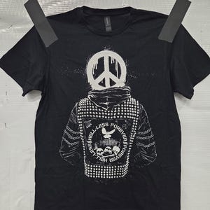 May include: Black t-shirt featuring a white graphic of a person wearing a peace sign helmet and a studded jacket. The jacket design includes a dove, skulls, and the text "Less Hell Forever".
