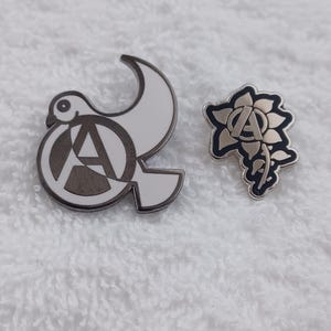 Könnte beinhalten: Zwei Metallnadeln. Eine Nadel ist eine weiße Taube mit einem grauen "A"-Symbol in der Mitte. Die andere Nadel ist ein graues "A"-Symbol in einem schwarz-silbernen Blumendesign. Beide Nadeln befinden sich auf einer weißen, strukturierten Oberfläche.