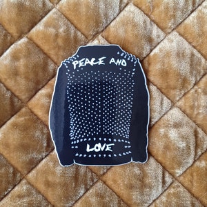 Op de afbeelding: Zwart-witte sticker van een leren jack met studs met de woorden "Peace and Love" erop.