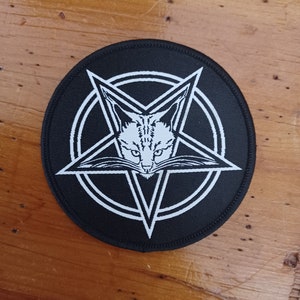 Pode incluir: Patch bordado em preto e branco com um pentagrama e um rosto de gato dentro. O patch é circular e tem um fundo preto.