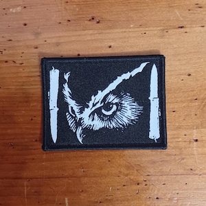 Puede incluir: Parche rectangular negro con un diseño de ojo de búho blanco. El ojo del búho está en el centro, flanqueado por dos barras verticales blancas. El parche es probablemente para ropa o accesorios.