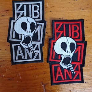 May include: Two embroidered patches featuring a skull design and the word "SUBHUMAN" in black lettering. The patch on the left has a white background and black border. The patch on the right has a red background and black border.