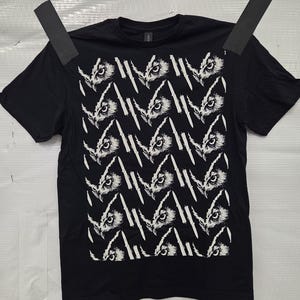 May include: Black t-shirt featuring a repeating pattern of white owl faces and diagonal lines. The design covers the front of the shirt. The shirt has short sleeves and a crew neck.
