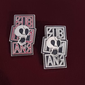 May include: Two black and white enamel pins with a skull design. The pins feature the text "SUBURBAN" and "ANZ" in a bold, stylized font. One pin has red accents, while the other has silver accents.