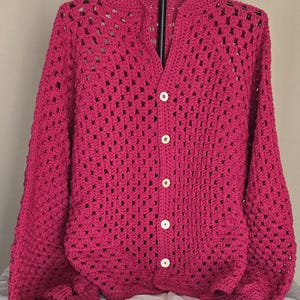 May include: A vibrant fuchsia long-sleeve cardigan featuring a classic granny square crochet pattern. The garment has a round neckline, ribbed cuffs, and a front button closure with five round white buttons. The open-knit texture is consistent throughout.