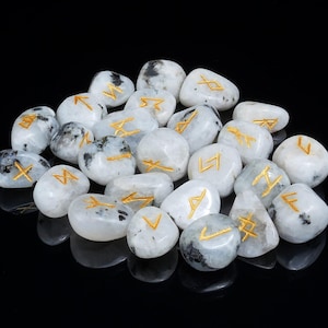 Peut inclure: Un ensemble de pierres de runes en pierre de lune blanche polie, avec des symboles gravés de couleur or. Les pierres varient en taille et en forme, certaines présentant des inclusions gris foncé. Les runes sont disposées sur une surface noire réfléchissante.