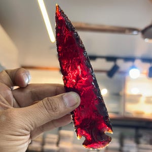 May include: A deep red obsidian arrowhead with a pointed tip and a jagged edge. The arrowhead is translucent and has a glossy finish.