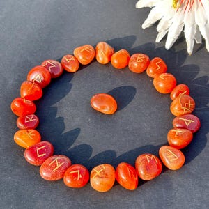 May include: A circular arrangement of orange-red stones, each etched with a golden symbol. A single stone rests in the centre. The stones are on a dark surface, with a white flower with a red centre in the background.