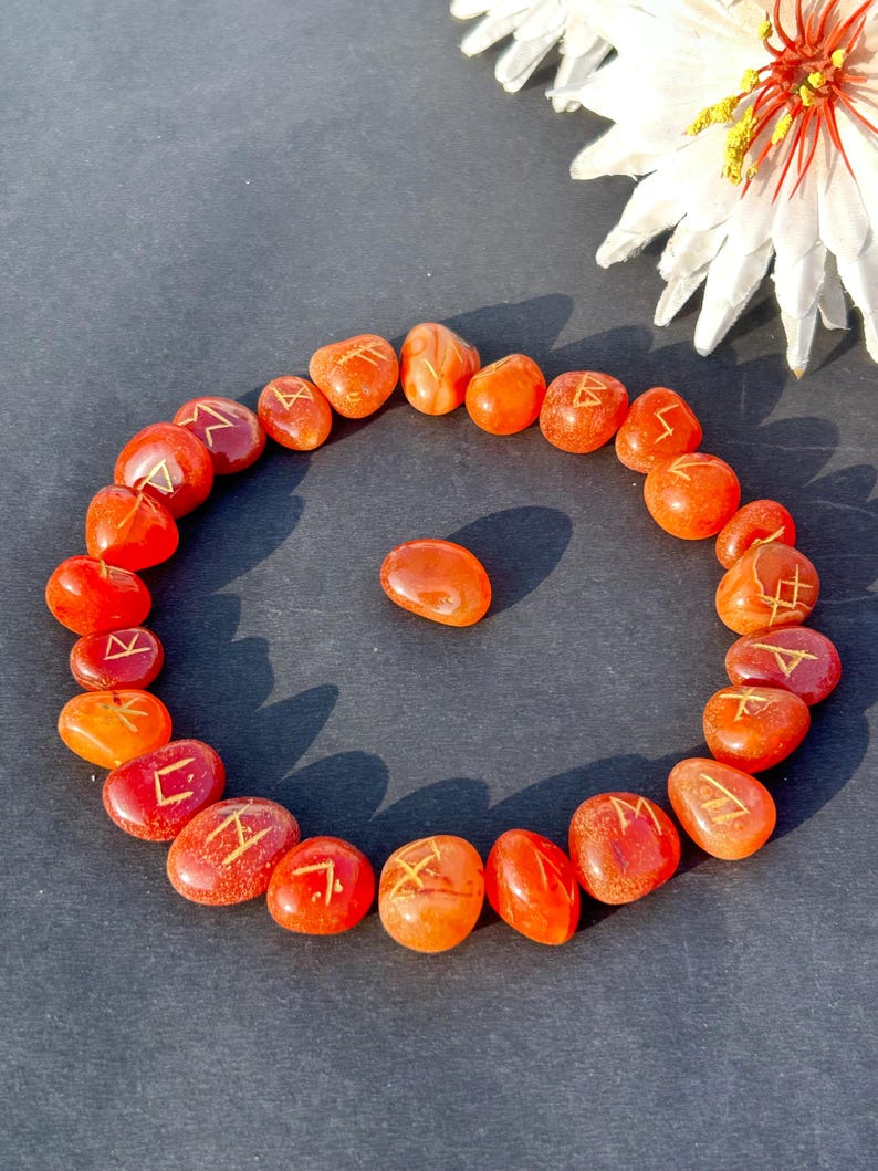 May include: A circular arrangement of orange-red stones, each etched with a golden symbol. A single stone rests in the centre. The stones are on a dark surface, with a white flower with a red centre in the background.