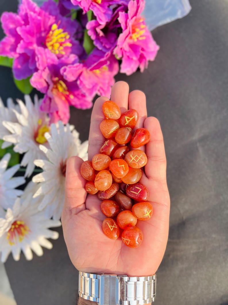May include: A collection of orange-red rune stones, each inscribed with gold symbols. The stones are held in a hand, and the background features purple and white flowers. The stones have a smooth, polished finish.