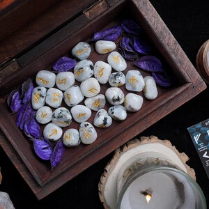 May include: A wooden box filled with white rune stones with gold lettering and purple decorative leaves. A lit candle, a wooden bowl, and a card with rune symbols are also visible. The scene is set on a dark surface.