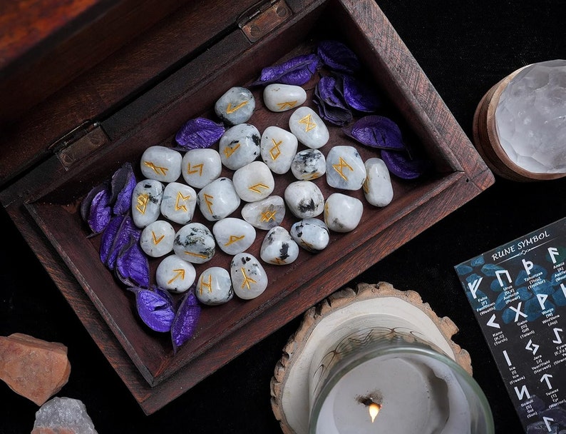 May include: A wooden box filled with white rune stones with gold lettering and purple decorative leaves. A lit candle, a wooden bowl, and a card with rune symbols are also visible. The scene is set on a dark surface.