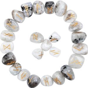 May include: A collection of white and gray moonstone runes with gold-colored etched symbols. The stones are arranged in a circular pattern, with some stones featuring black inclusions. The runes are likely used for divination or spiritual practices.