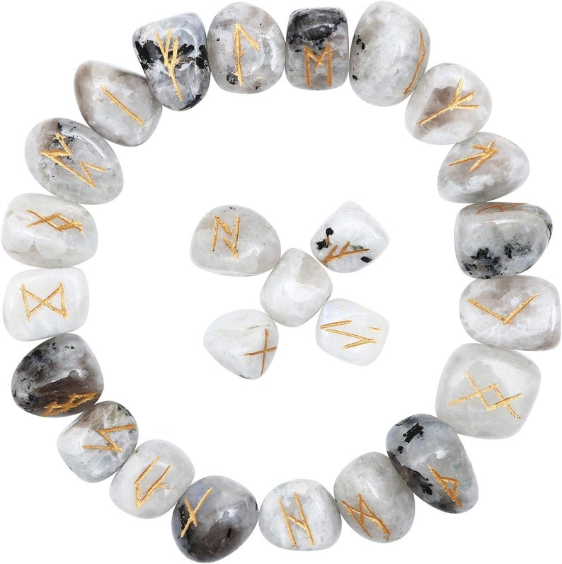 May include: A collection of white and gray moonstone runes with gold-colored etched symbols. The stones are arranged in a circular pattern, with some stones featuring black inclusions. The runes are likely used for divination or spiritual practices.