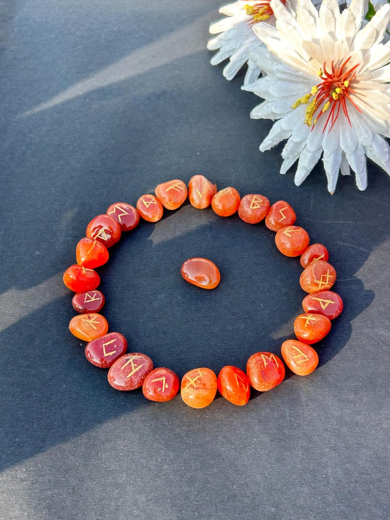 May include: A ring of orange and red rune stones, each inscribed with gold symbols. A single stone sits in the centre. A white flower with a yellow centre is in the background. The stones are on a dark surface.