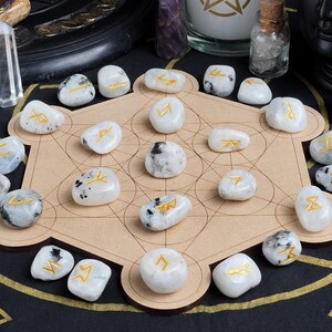 May include: A collection of white moonstone rune stones with gold symbols arranged on a wooden Metatron's Cube. The stones are surrounded by other spiritual items, including crystals and candles, on a black surface.