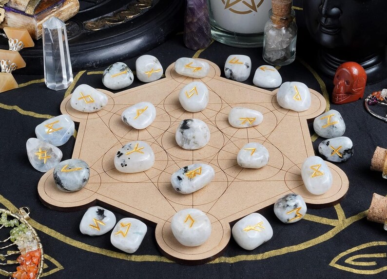 May include: A collection of white moonstone rune stones with gold symbols arranged on a wooden Metatron's Cube. The stones are surrounded by other spiritual items, including crystals and candles, on a black surface.