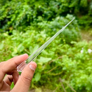 Puede incluir: Una herramienta de vidrio transparente y puntiaguda, sostenida contra un fondo verde borroso. La herramienta tiene una punta larga y afilada y un mango rectangular. El objeto se sostiene en una mano, con el foco en la herramienta de vidrio.