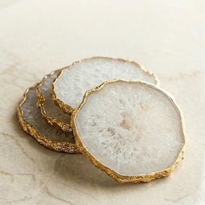 May include: Four white agate coasters with gold gilded edges. The coasters have a natural, organic shape and a translucent appearance. The gold edges add a touch of elegance to the coasters, which are suitable for home decor.