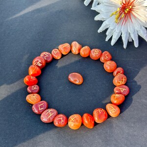 May include: A ring of orange and red rune stones, each inscribed with gold symbols. A single stone sits in the centre. A white flower with a yellow centre is in the background. The stones are on a dark surface.