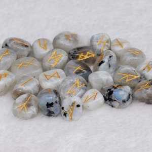 May include: A collection of polished, oval-shaped moonstone runes. Each stone is a mix of white, gray, and black with gold-colored etched symbols. The runes are arranged on a soft, white surface.